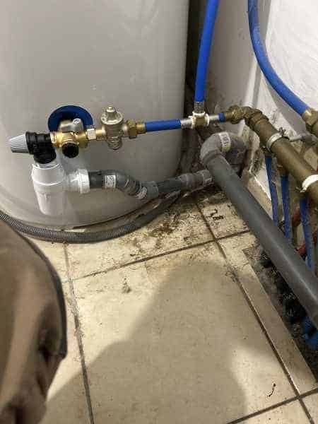 Raccordements de plomberie en laiton et tuyaux bleus fixés à un chauffe-eau cylindrique, près du sol carrelé sale.