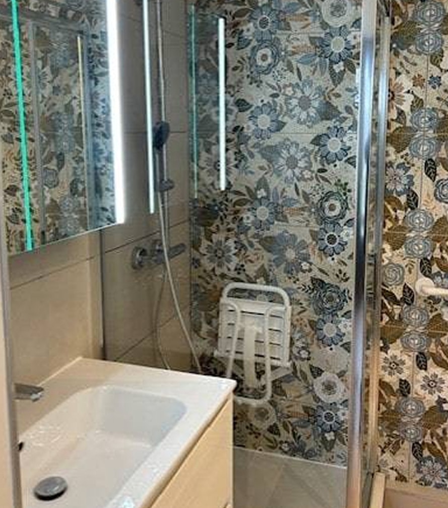 Douche moderne carrelage floral et siège PMR Salle de bain moderne avec lavabo et douche vitrée. Carrelage mural floral bleu et marron. Siège de douche mural pliant blanc.