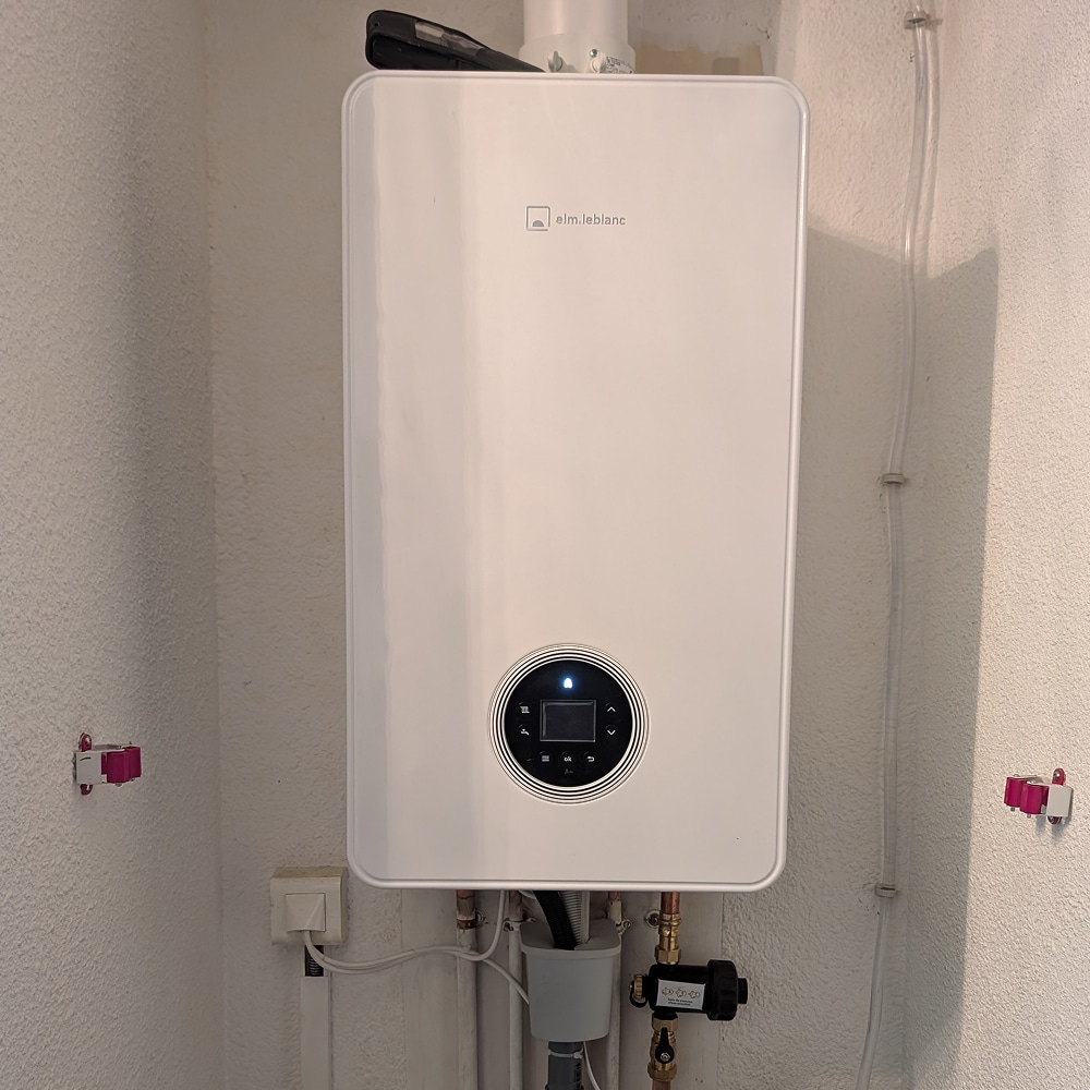 Chaudière murale gaz Elm.leblanc, modèle blanc avec interface digitale ronde et connexions de plomberie visibles en dessous.