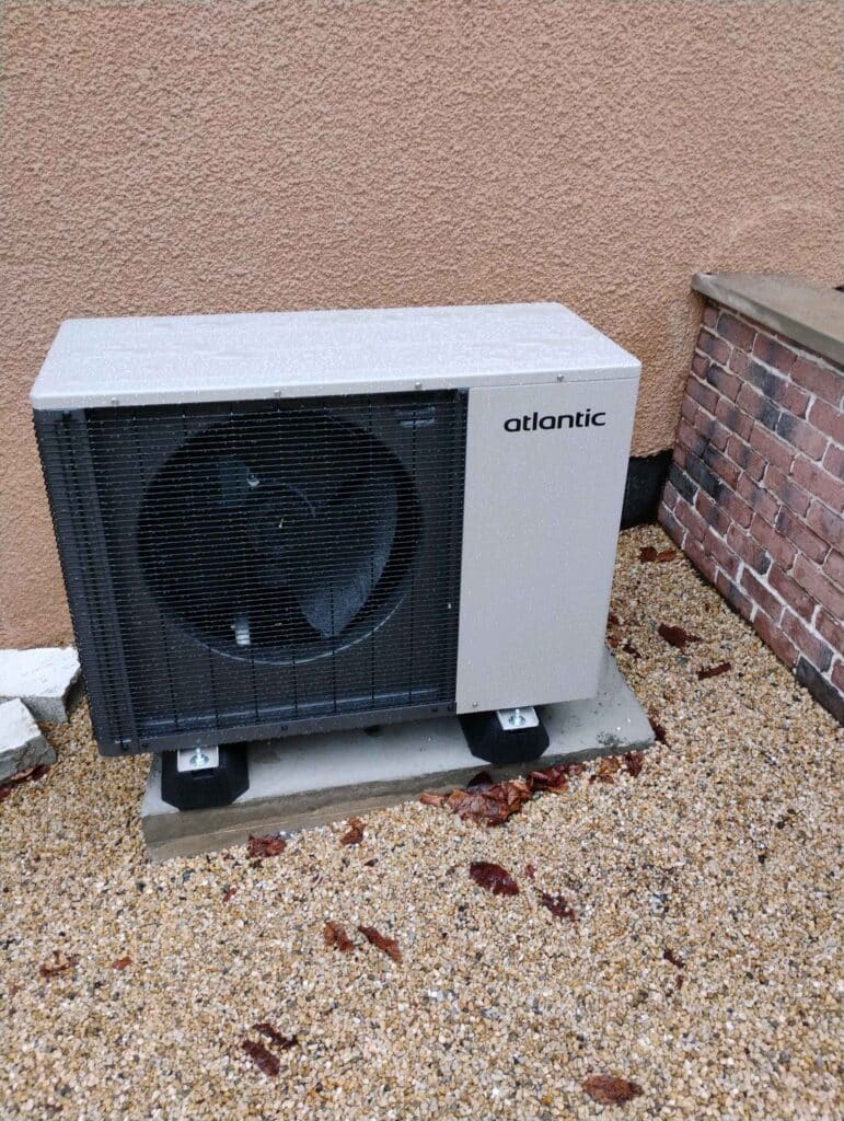 Unité extérieure de pompe à chaleur Atlantic, grise et noire, installée sur une dalle de béton au-dessus de gravier, sous la pluie.