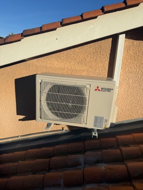 Unité extérieure de climatisation Mitsubishi Electric fixée sur un support mural au-dessus d'un toit de tuiles rouges sous un ciel bleu clair.