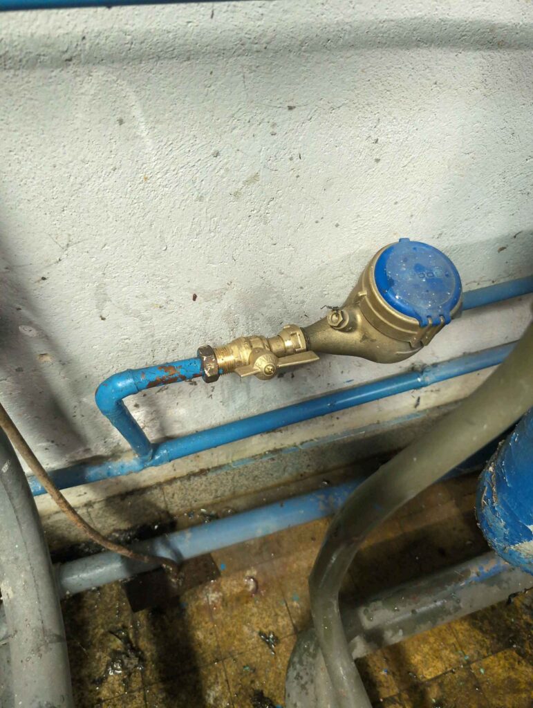 Compteur d'eau en laiton à capot bleu, installé sur un tuyau de plomberie bleu. Une vanne d'arrêt "RAVA" est visible en amont.