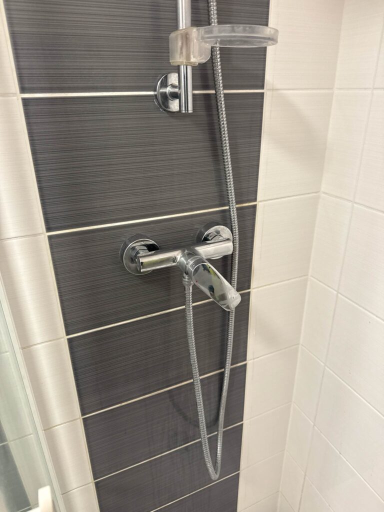 Mitigeur de douche chromé sur carrelage moderne gris foncé à rayures horizontales, encadré par des carreaux blancs clairs. Flexible et support visible.