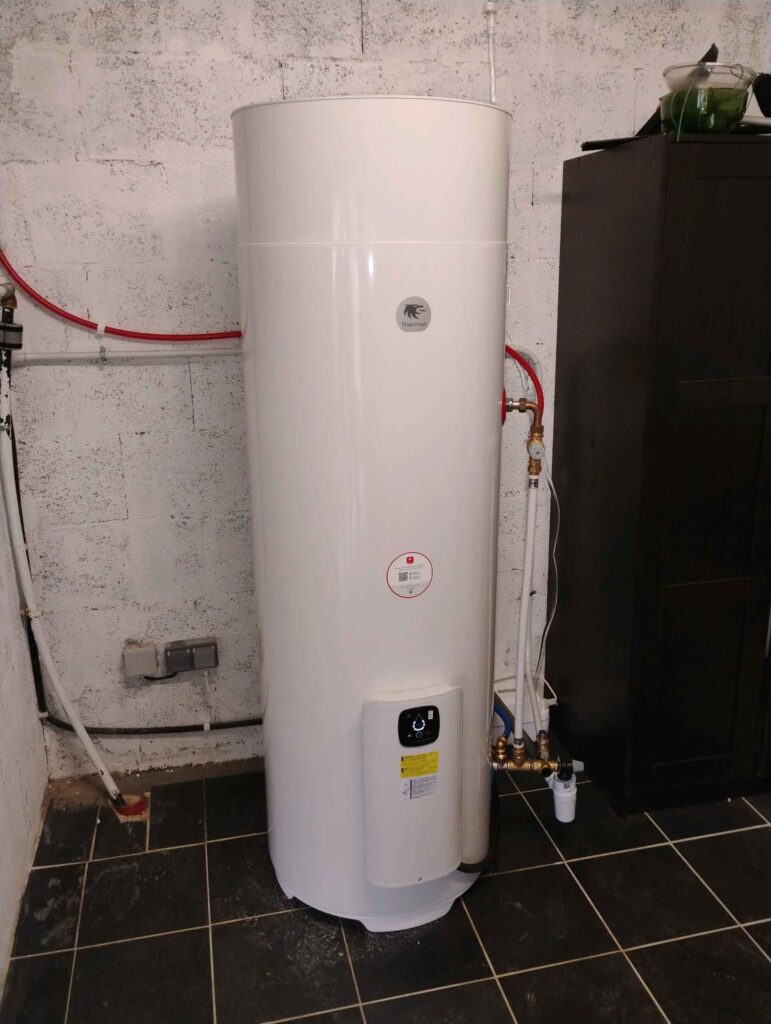 Chauffe-eau thermodynamique blanc Thermor installé avec tuyauterie apparente dans une buanderie.