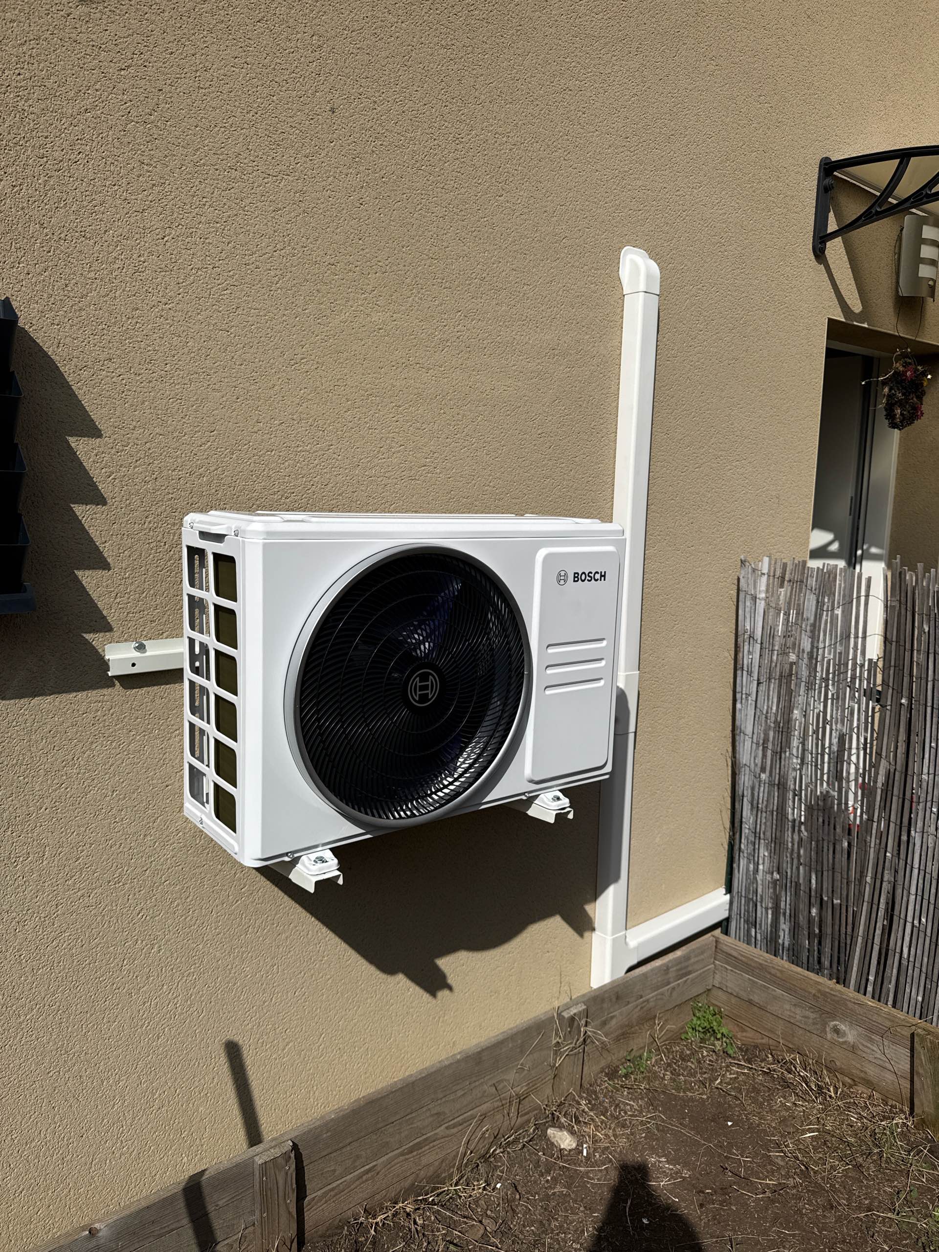 Unité extérieure de climatisation Bosch blanche fixée sur un mur beige avec supports et goulotte de protection.