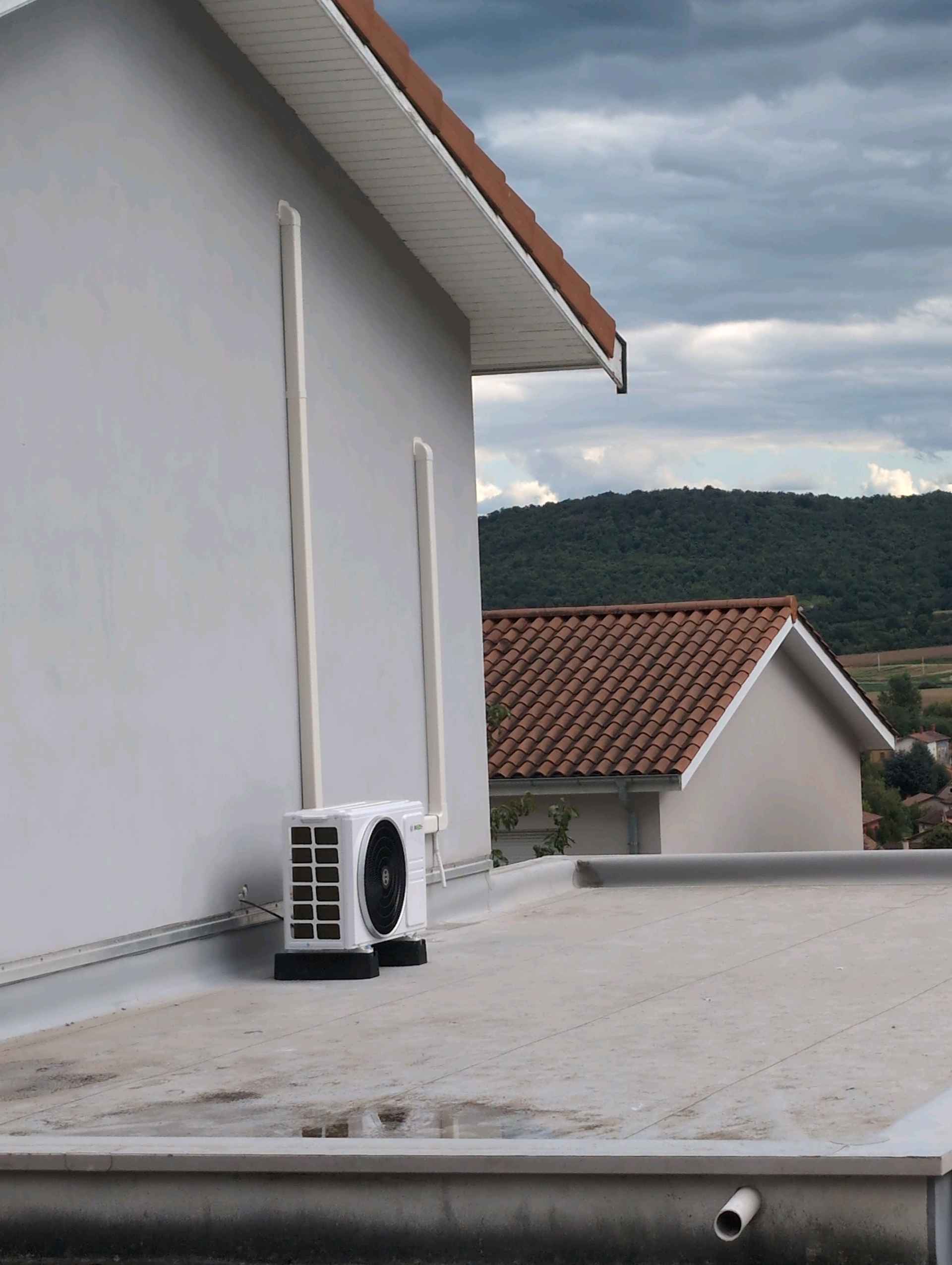 Unité extérieure de climatisation blanche sur un toit plat avec conduits verticaux devant un paysage de collines. vienne