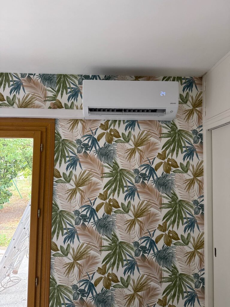 Climatiseur mural Atlantic affichant 24°C sur un papier peint aux motifs de feuilles tropicales colorées.